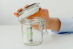 Aperson opening a glass jar with money inside it.